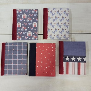 May include: Five rectangular notebooks with fabric spines. The top left features a farm pattern, top right has donkeys in hats, bottom left is a blue grid, bottom center is red with white stars, and bottom right displays a stars and stripes design.