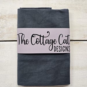 Puede incluir: Tela gris oscuro doblada con una banda lila. La banda presenta el texto "The Cottage Cat DESIGNS" en negro, con una fuente cursiva. La tela está sobre una superficie de madera blanca.