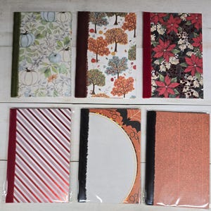 May include: Six rectangular notebooks with varied seasonal covers. Top row: pumpkins on white, autumn trees, and red poinsettias. Bottom row: red and white diagonal stripes, a Halloween bat design, and a vintage-style orange text pattern.