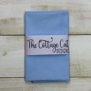 May include: A folded, light blue fabric with a textured surface. A white band wrapped around the fabric displays the text "The Cottage Cat Designs" in black script. The fabric is set against a light wood grain background.