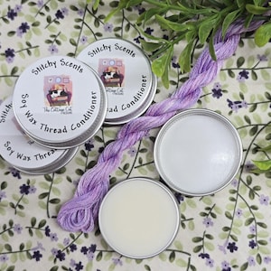 May include: Four small round tins of soy wax thread conditioner, labeled "Stitchy Scents" and "The Cottage Cat Creations". The tins are on a floral fabric background with a spool of purple embroidery floss.