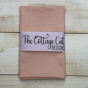 May include: A folded, blush-colored fabric with a textured appearance. A white band wrapped around the fabric displays the text "The Cottage Cat DESIGNS" in black script. The fabric is resting on a light-colored wooden surface.