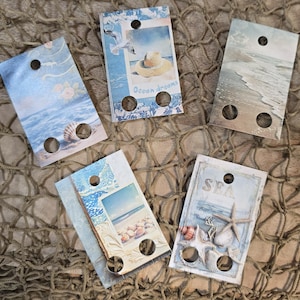 May include: Five rectangular tags with beach-themed designs. Each tag features a different seaside image, including seashells, waves, and a seagull. The tags have a hole at the top and two at the bottom.