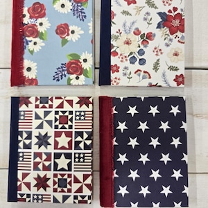 May include: Four rectangular journals with fabric-covered hardcovers. The top left features red roses on light blue with a red fringe spine. The top right has a floral print on white with a navy spine. The bottom left shows a red, white, and blue quilt pattern with a navy spine. The bottom right displays white stars on navy with a red fringe spine.