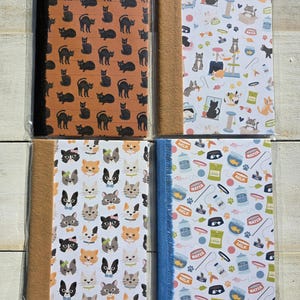 May include: Four rectangular notebooks with cat-themed covers. One has black cats on orange, one has cat toys and accessories on white, one has various cat faces on white, and one has milk cartons and pet supplies on white with a blue fabric spine.