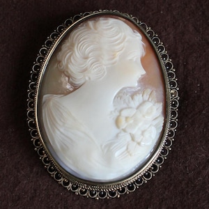 May include: Oval cameo brooch depicting a woman's profile with curly hair and a floral bouquet. The cameo is set in a gold-toned frame with a decorative border of small black beads. The background is a warm, light brown.