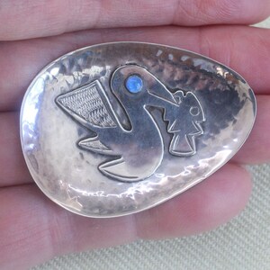 Puede incluir: Un colgante de plata con forma de pájaro con una piedra azul para el ojo. El pájaro sostiene un pequeño árbol en su pico. El colgante está martillado y tiene una superficie texturizada.