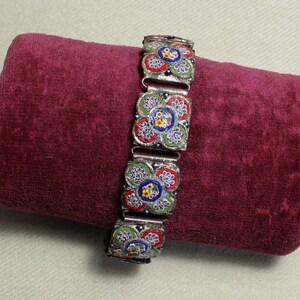 May include: A silver bracelet with a floral design made of colorful mosaic tiles. The bracelet is on a red velvet surface.