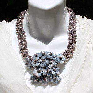 May include: A statement necklace featuring a large cluster of gray and white stone flowers with a delicate chain of small, iridescent beads.