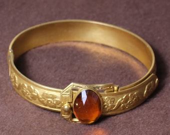 May include: A gold-toned bangle bracelet with an oval-shaped amber-colored glass stone set in a decorative metal frame. The bracelet is decorated with floral designs.