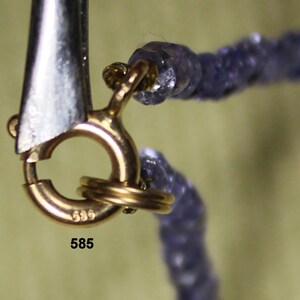 May include: Close-up of a gold clasp with the number 585 stamped on it. The clasp is attached to a necklace made of small, purple beads.