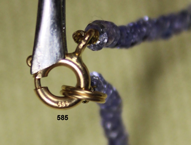 May include: Close-up of a gold clasp with the number 585 stamped on it. The clasp is attached to a necklace made of small, purple beads.