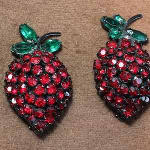 May include: A pair of red and green rhinestone strawberry brooches. The strawberries are made of red rhinestones and have green rhinestone leaves.