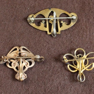 May include: Three gold-toned brooches with intricate designs. The brooches are on a brown fabric background.