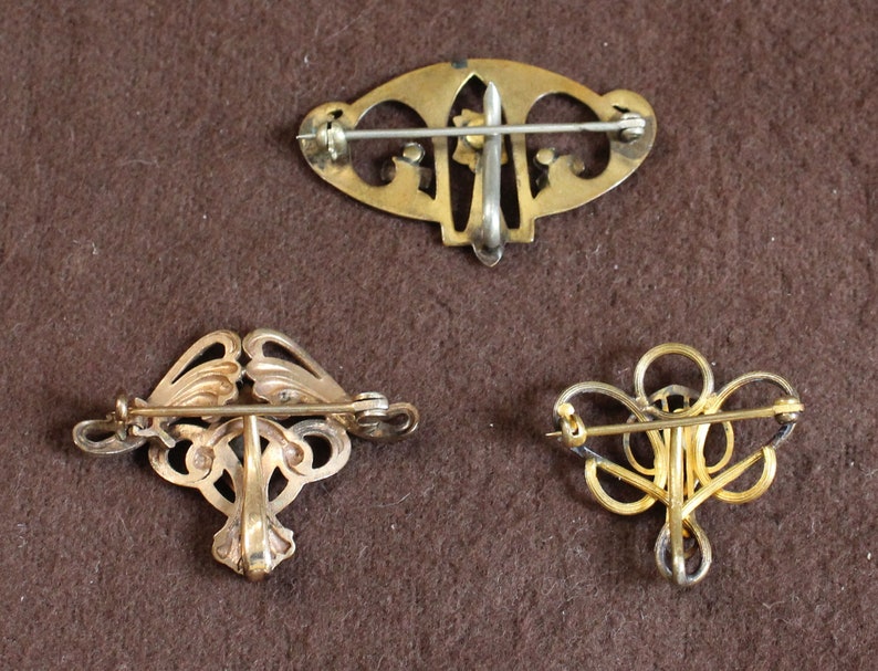 May include: Three gold-toned brooches with intricate designs. The brooches are on a brown fabric background.