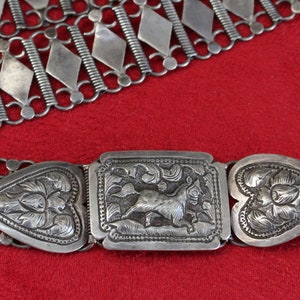 May include: A silver belt with an ornate buckle featuring a stylized animal design. The belt is made of linked silver chains with diamond-shaped links.
