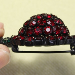 May include: A vintage brooch with a cluster of red rhinestones set in a black metal base. The brooch has a pin and catch closure.