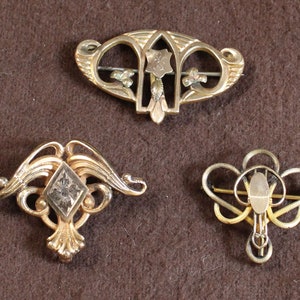 May include: Three gold-toned brooches with intricate designs. The top brooch features a floral motif with a central shield. The left brooch has a diamond-shaped center with a floral design. The right brooch has a heart-shaped center with a floral design.