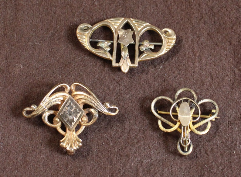 May include: Three gold-toned brooches with intricate designs. The top brooch features a floral motif with a central shield. The left brooch has a diamond-shaped center with a floral design. The right brooch has a heart-shaped center with a floral design.