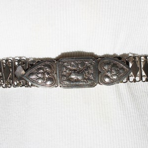 May include: A silver chain belt with a heart-shaped buckle and a central rectangular buckle with an ornate design.