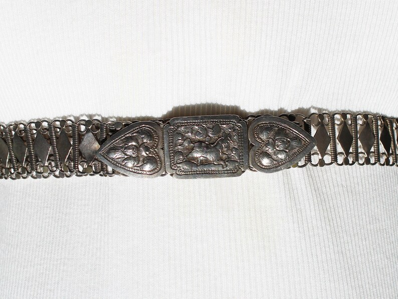 May include: A silver chain belt with a heart-shaped buckle and a central rectangular buckle with an ornate design.