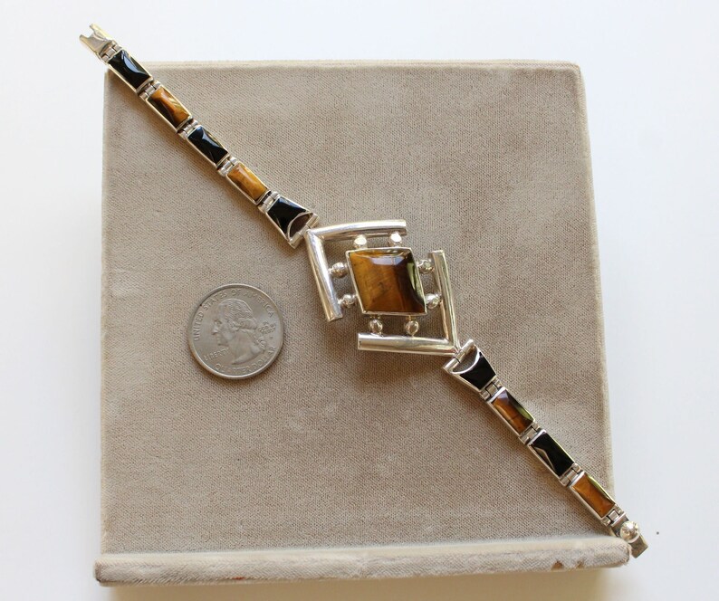 May include: A silver bracelet with a geometric design featuring a large tiger's eye stone. The bracelet is on a brown velvet surface.