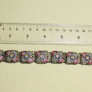 May include: A silver bracelet with a series of square-shaped mosaic tiles. The tiles are decorated with a floral pattern in shades of green, red, blue, and white.