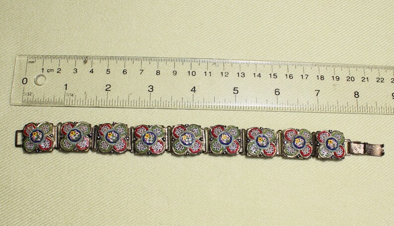 May include: A silver bracelet with a series of square-shaped mosaic tiles. The tiles are decorated with a floral pattern in shades of green, red, blue, and white.