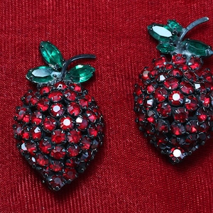 May include: A pair of red rhinestone earrings shaped like strawberries with green rhinestone leaves.