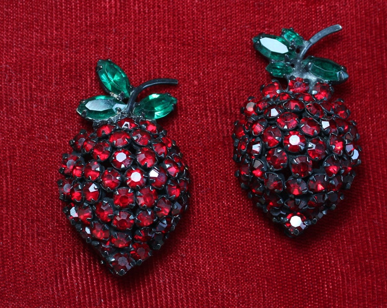 May include: A pair of red rhinestone earrings shaped like strawberries with green rhinestone leaves.