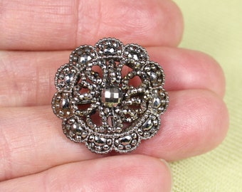 May include: A silver-toned, round, decorative button with a flower-like design made of small, faceted stones. The center of the button features a larger, faceted stone.
