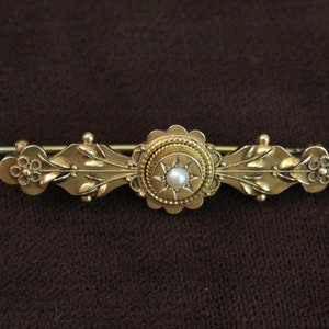 Puede incluir: Un broche de oro con una perla en el centro, rodeada de un diseño de estrella. El broche está decorado con motivos florales.