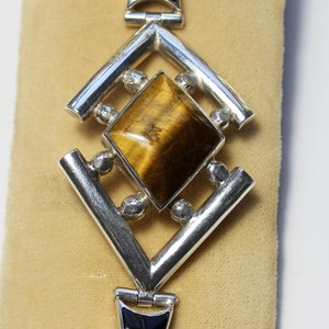 May include: A silver bracelet with a geometric design featuring a large, square-shaped tiger's eye gemstone.