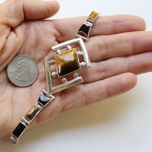 May include: A silver bracelet with a square tiger's eye gemstone in the center. The bracelet is made of silver and has black accents.