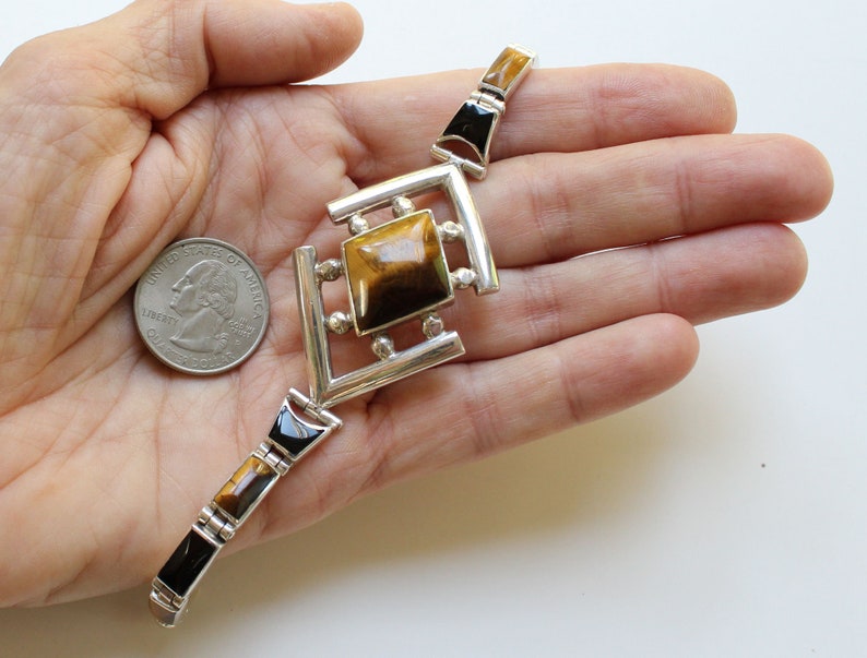 May include: A silver bracelet with a square tiger's eye gemstone in the center. The bracelet is made of silver and has black accents.