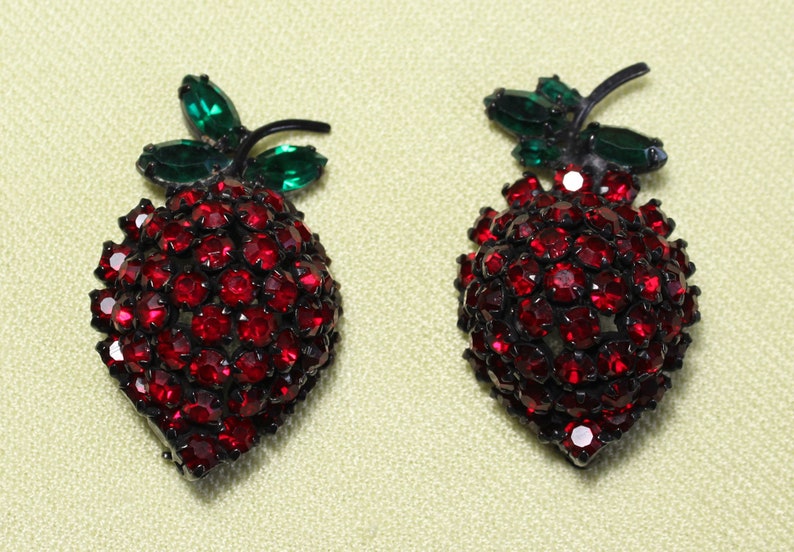 May include: A pair of red rhinestone strawberry brooches with green rhinestone leaves.