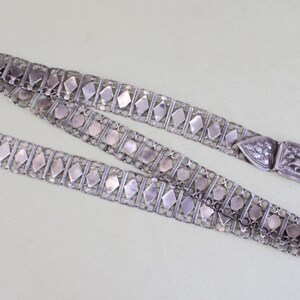 May include: A silver chain belt with a decorative clasp. The chain is made of interlocking links with a diamond-shaped pattern. The clasp is ornate and features a floral design.