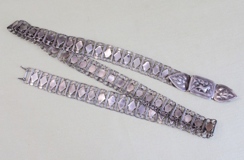 May include: A silver chain belt with a decorative clasp. The chain is made of interlocking links with a diamond-shaped pattern. The clasp is ornate and features a floral design.