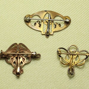 May include: Three gold-toned brooches with intricate filigree designs. The brooches are all different shapes and sizes, and they have a vintage look.