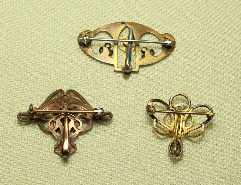 May include: Three gold-toned brooches with intricate filigree designs. The brooches are all different shapes and sizes, and they have a vintage look.