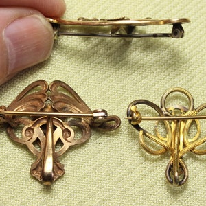 May include: Three gold-toned brooches with intricate designs. The brooches are made of metal and have a delicate, filigree-like appearance. They are all different shapes and sizes, but they share a similar style.
