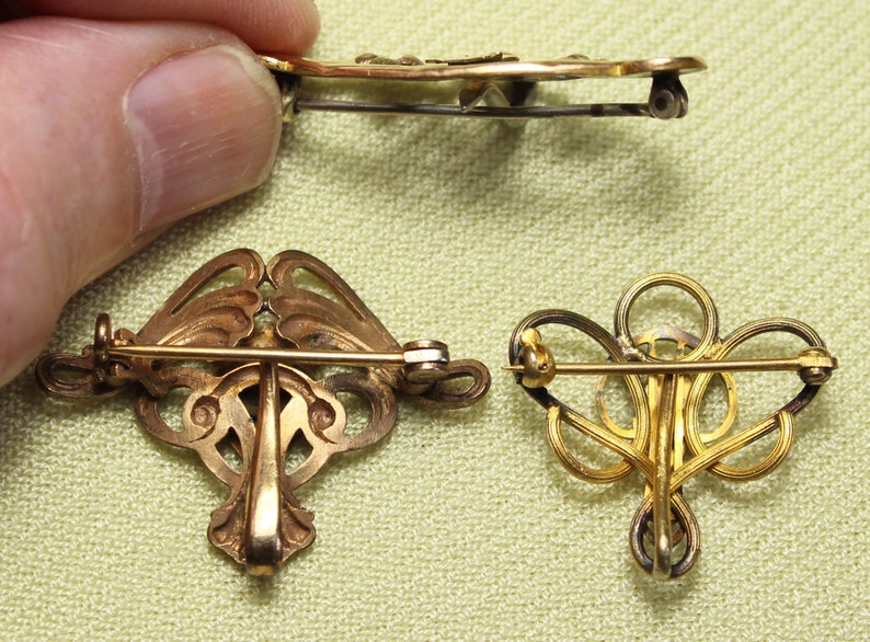 May include: Three gold-toned brooches with intricate designs. The brooches are made of metal and have a delicate, filigree-like appearance. They are all different shapes and sizes, but they share a similar style.