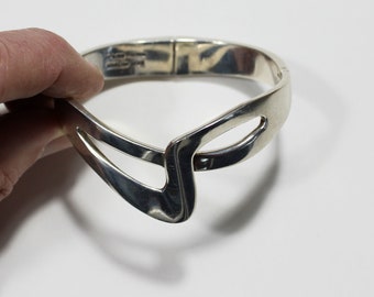 May include: A silver bangle bracelet with a unique, abstract design. The bracelet is open and has a hinge closure.