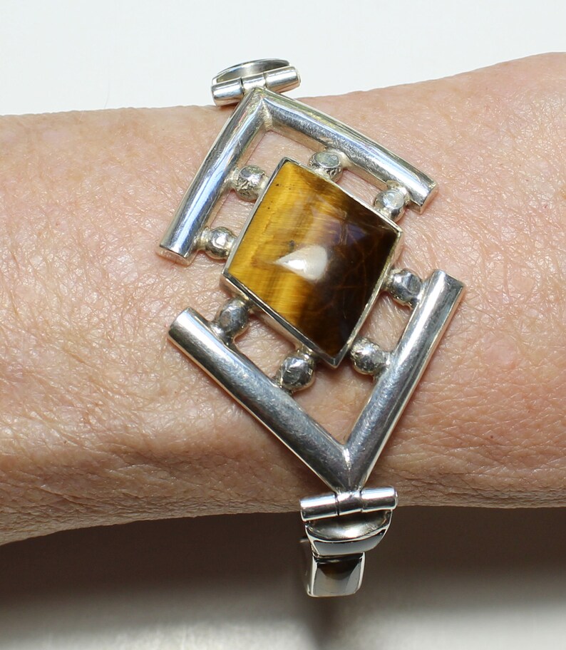 May include: A silver bracelet with a geometric design featuring a large, rectangular tiger's eye gemstone.