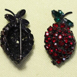 May include: Two vintage brooch pins in the shape of a bunch of grapes. One is black with red rhinestones and the other is red with green leaves.