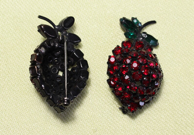 May include: Two vintage brooch pins in the shape of a bunch of grapes. One is black with red rhinestones and the other is red with green leaves.