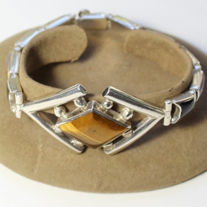 May include: Silver bracelet with a large tiger's eye gemstone in a geometric setting. The bracelet is made of linked silver bars with black onyx accents.