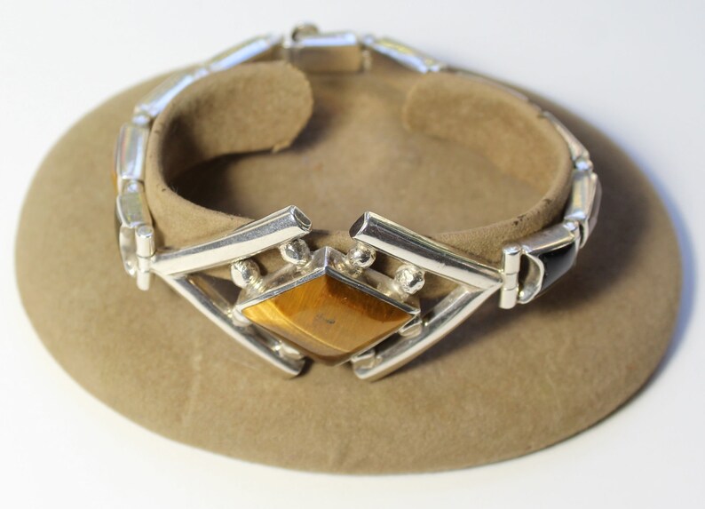 May include: Silver bracelet with a large tiger's eye gemstone in a geometric setting. The bracelet is made of linked silver bars with black onyx accents.