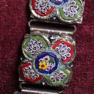 May include: A close-up of a silver bracelet with a floral design made of colorful mosaic tiles. The tiles are arranged in a pattern of flowers and swirls, with shades of red, white, blue, green, and yellow.