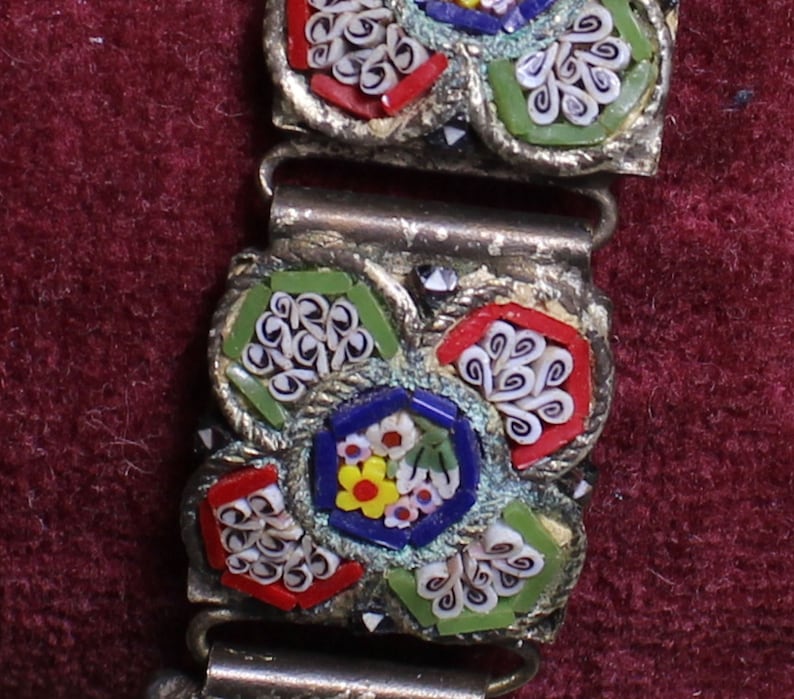 May include: A close-up of a silver bracelet with a floral design made of colorful mosaic tiles. The tiles are arranged in a pattern of flowers and swirls, with shades of red, white, blue, green, and yellow.